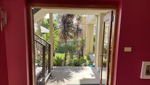 L’entrée d’un hôtel avec un mur rouge, une porte en verre, un paillasson noir et une vue sur un jardin planté de palmiers et un bâtiment en briques.