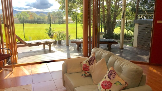 Une pièce avec vue sur un champ verdoyant et des arbres, une terrasse en bois à l’extérieur et un canapé beige agrémenté de coussins à motifs floraux.