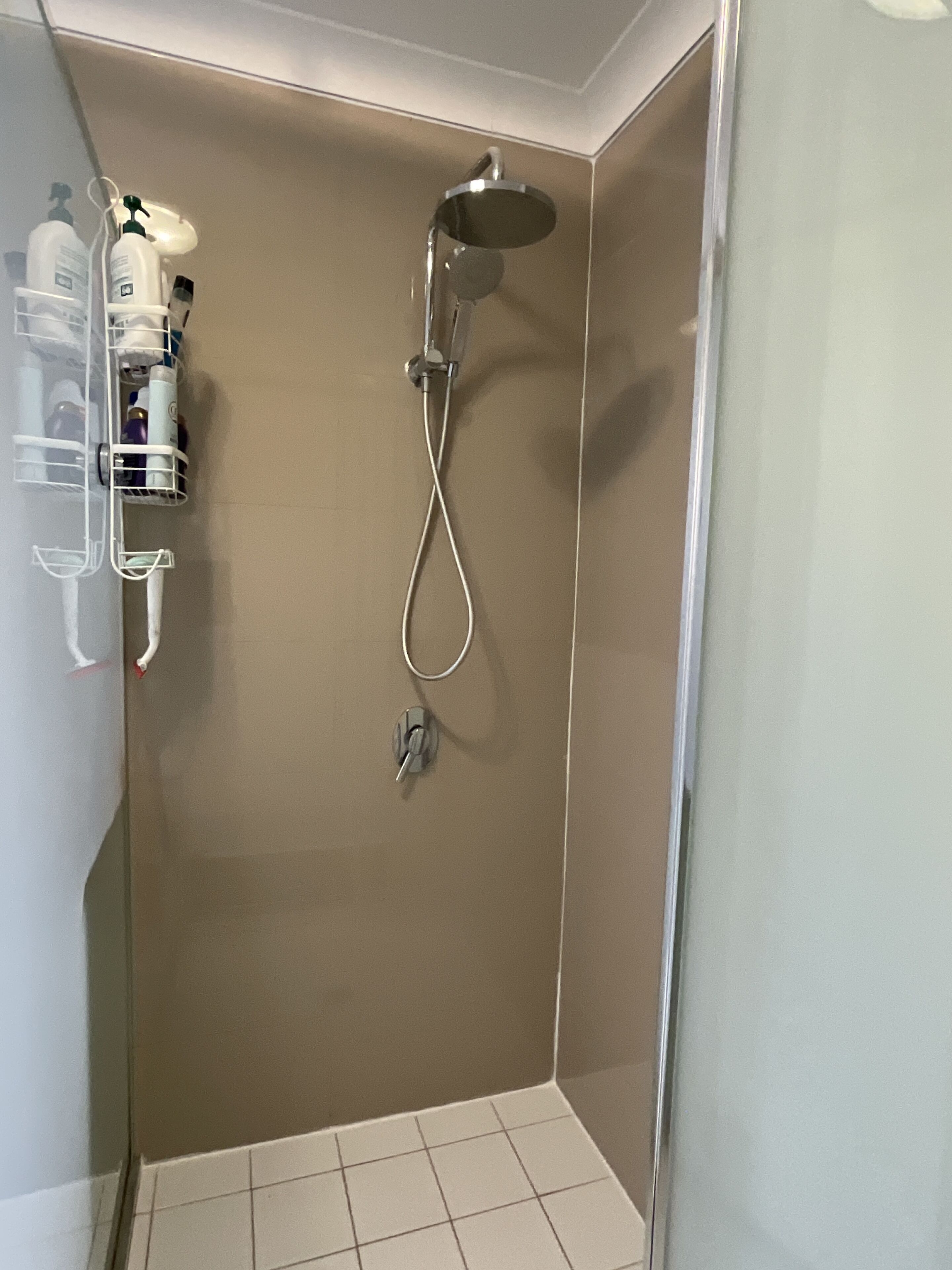 Un petit espace douche fermé, doté d’un pommeau de douche et d’une manette de réglage, d’une étagère pour les articles de toilette et d’un sol carrelé.