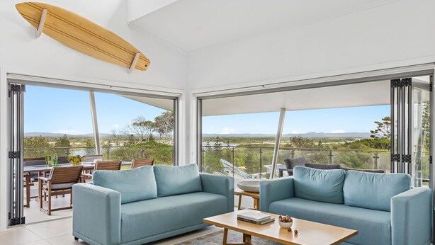 A modern living room with two sofas, a coffee table, and a surfboard on the ceiling. Large windows offer a view of the outdoors. - Pilotage 4 - Luxury Ocean and River View Penthouse - Holiday Yamba (Yamba)