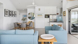 A modern living room with light blue sofas, a dining area with a wooden table, and a kitchen with white cabinets and stainless steel appliances. - Pilotage 4 - Luxury Ocean and River View Penthouse - Holiday Yamba (Yamba)