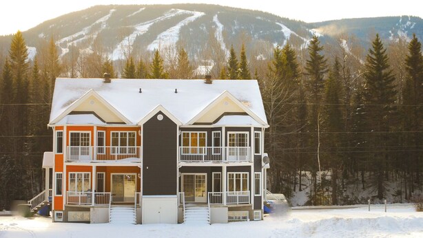 Une maison à deux étages, avec une partie centrale de couleur sombre et des murs extérieurs orange et blancs, se dressant sur fond de montagnes enneigées.
