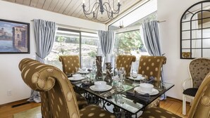 Uma área de jantar com mesa de vidro posta para quatro pessoas, rodeada por cadeiras estampadas, um lustre e grandes janelas com cortinas.