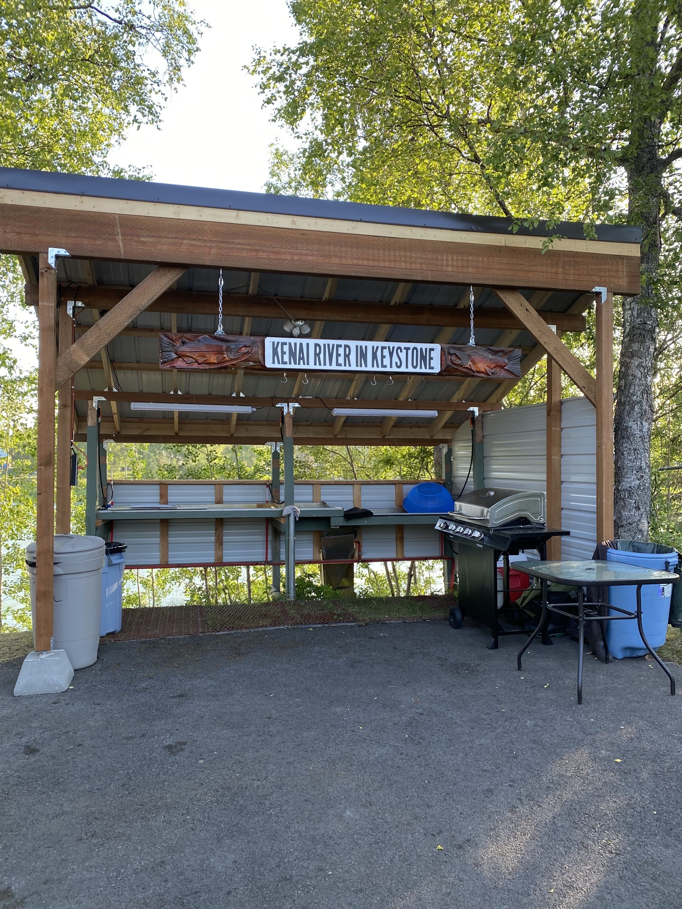 A covered picnic area with a sign reading "Kenai River in Keystone," a grill, and trash bins.