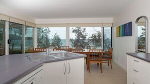 A modern kitchen with a large island, a dining area with wooden chairs, and a view of the sea through large windows. - Beachfront Duplex on Denhams (Denhams Beach)