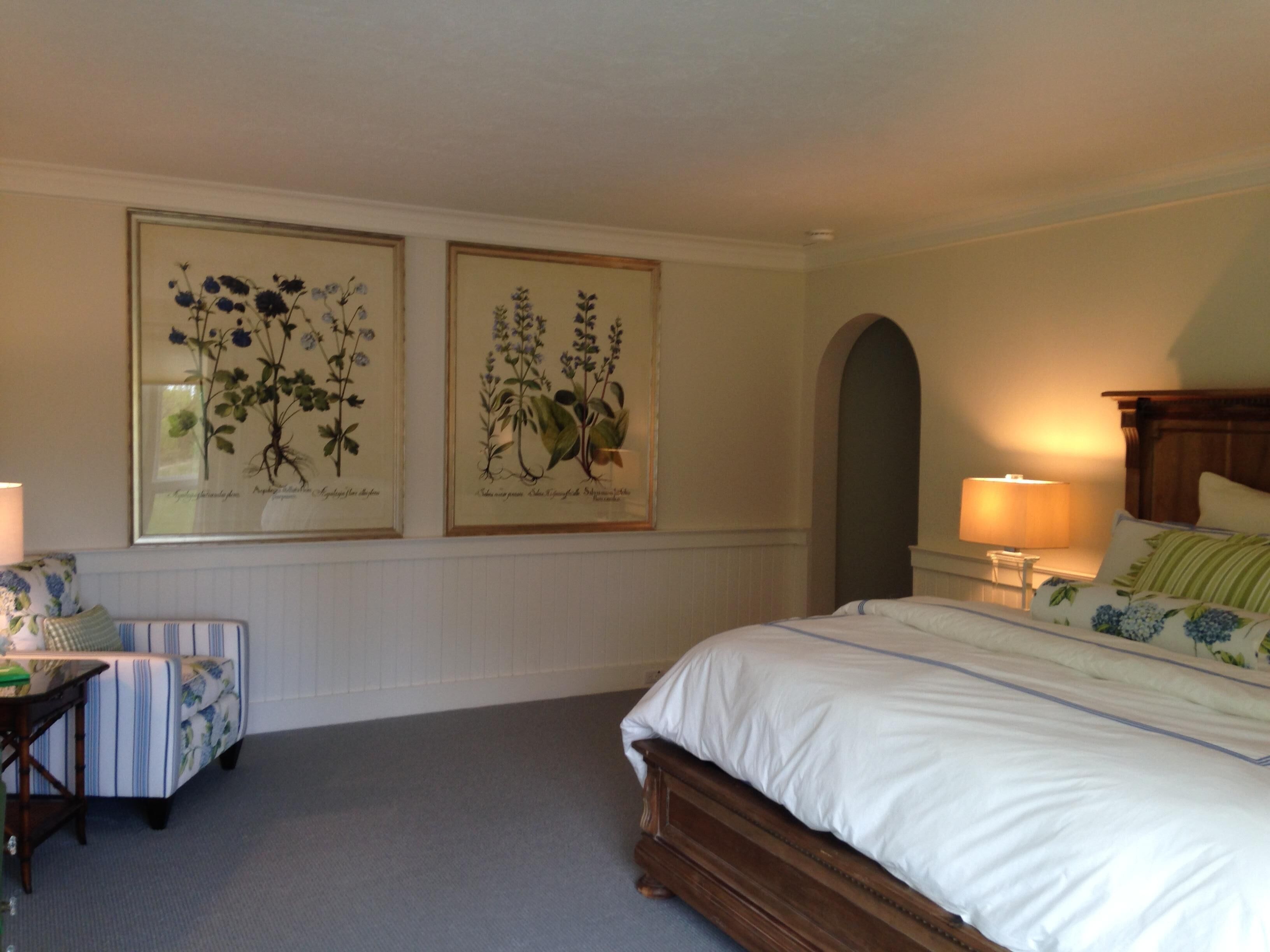 A bedroom with a bed, a nightstand, a chair, and two framed botanical illustrations on the wall.