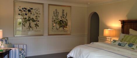 A bedroom with a bed, a nightstand, a chair, and two framed botanical illustrations on the wall.