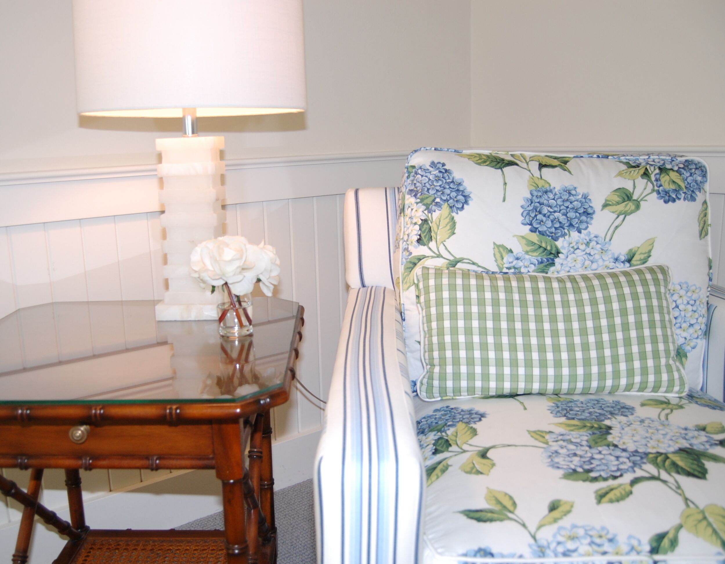 A room with a wooden table, a glass-top coffee table, a chair with floral upholstery, and a lamp.