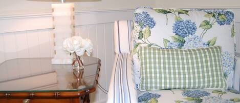 A room with a wooden table, a glass-top coffee table, a chair with floral upholstery, and a lamp.