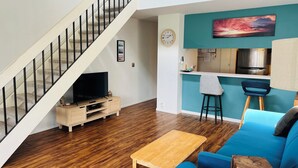 Una sala de estar moderna con piso de madera, un sofá azul, una mesa de centro de madera, un televisor sobre un mueble de madera, una escalera con barandillas negras, una zona de cocina con refrigerador de acero inoxidable y una pared de color verde azulado con una pintura de atardecer.