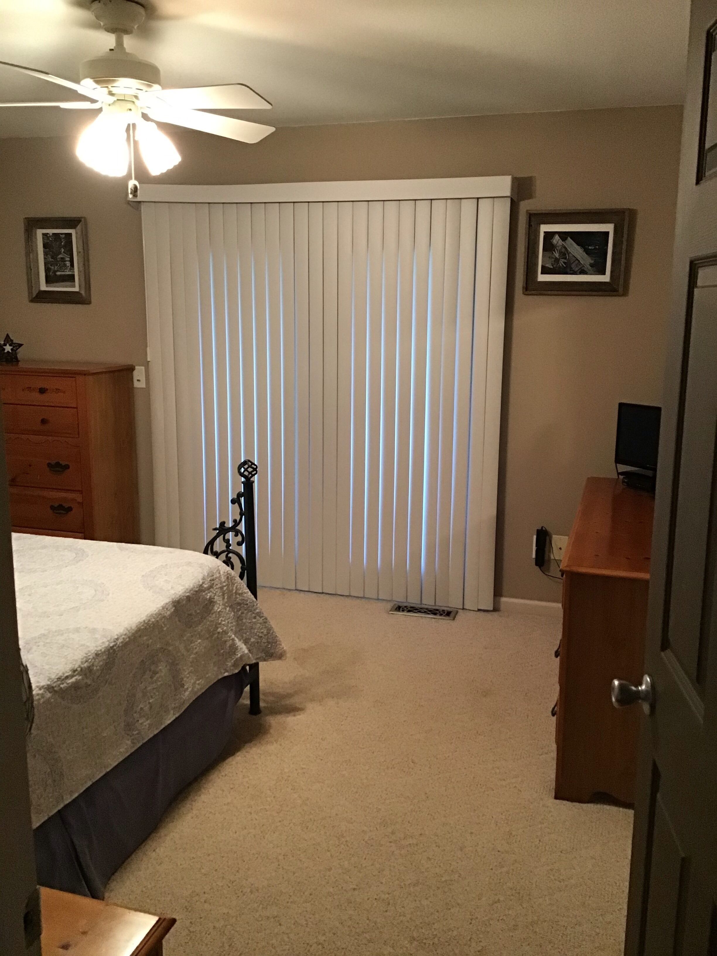 Ein Schlafzimmer mit Bett, Kommode, Schreibtisch, Deckenventilator und Fenster mit Jalousien.