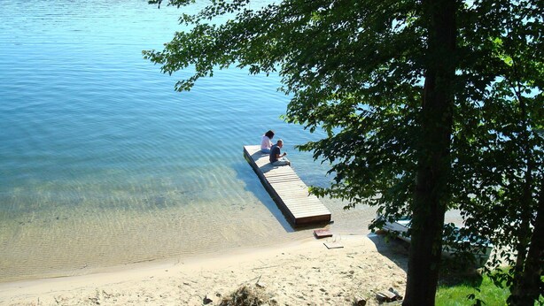 A wooden dock extending into a clear lake with two people sitting at the end.