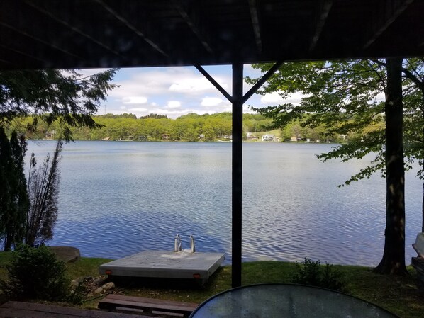 A lakeside view from a covered deck with a dock, surrounded by trees and houses in the background.