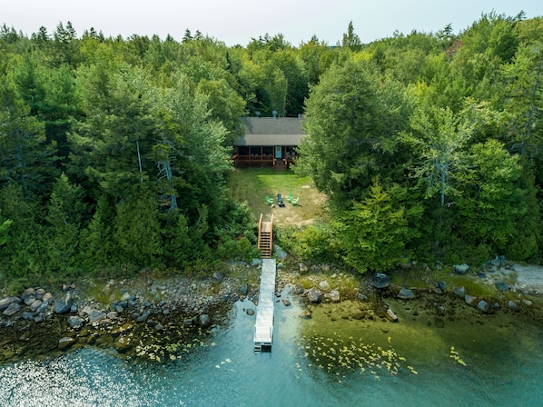 Une cabane entourée d’une forêt dense, un quai menant à une petite jetée, et une étendue d’eau claire parsemée de rochers et de nénuphars.