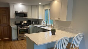 A modern kitchen with white cabinets, stainless steel appliances, a marble countertop, and a small island with two bar stools. - Family Cottage Close to Beach!! (Harwichport)