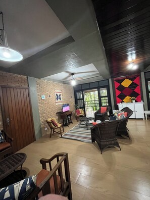 A spacious living area with a brick wall, a ceiling fan, a television, and a large window with a view of greenery. - Domingas Dias, 80m from the sea, 4 suites with pool. (Ubatuba)