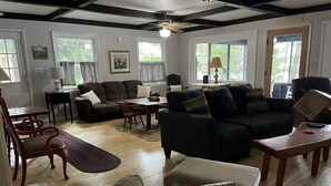 A cozy living room with a wooden floor, a brown sofa, a black sofa, a dining table, and a coffee table. There are two windows with curtains, a ceiling fan, and a lamp on a side table. - Picturesque Adirondack Lake Home (Greig)