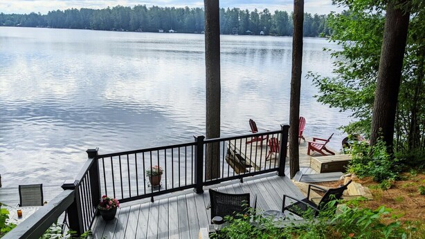 A wooden deck with a railing overlooking a calm lake, surrounded by trees. - Picturesque Adirondack Lake Home (Greig)