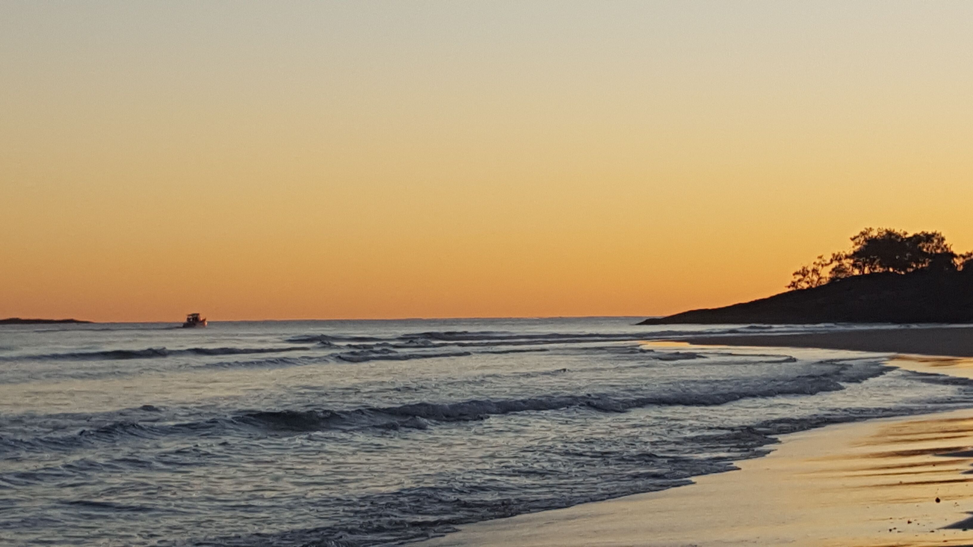 Een strand met golven bij zonsondergang, een boot in de verte en een heuvel met bomen.