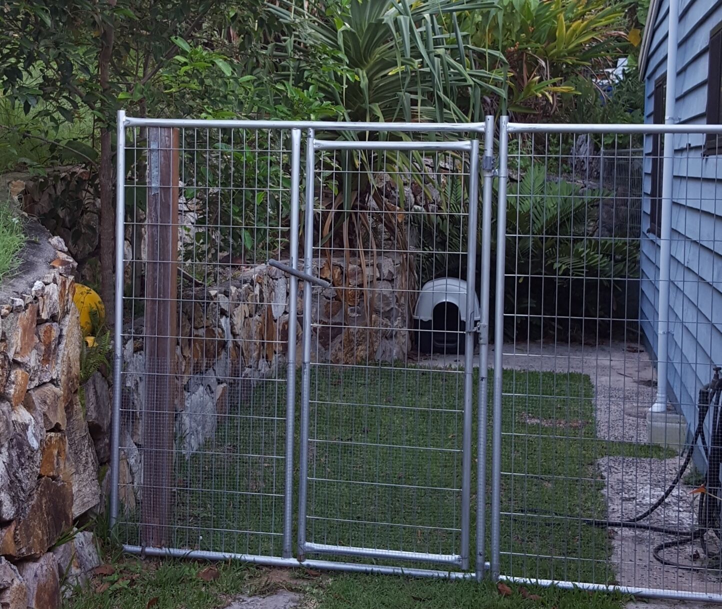 Ein Metalltor mit Drahtzaun, eine Steinmauer und ein kleines Tiergehege im Hintergrund.