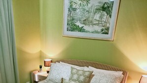 Una cama bien hecha con una colcha estampada, almohadas y toallas enrolladas. Hay un cuadro en la pared con una escena tropical, y las paredes son verdes.