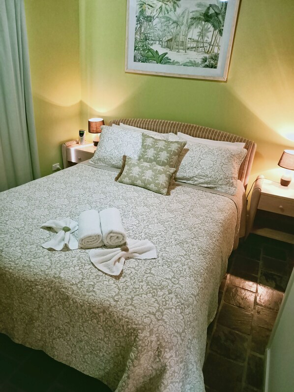Una cama bien hecha con una colcha estampada, almohadas y toallas enrolladas. Hay un cuadro en la pared con una escena tropical, y las paredes son verdes.