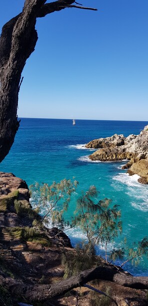 Un paisaje costero con terreno rocoso, mar azul cristalino y un velero a lo lejos.