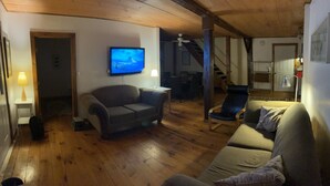 A cozy living room with a wooden floor, a grey sofa, a blue armchair, and a television on the wall. - Cooperstown Area Charming Carriage House (Mount Vision)