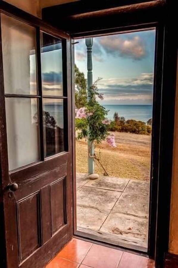 Una puerta de madera que da a un balcón con vistas al mar y una planta floreciente de color rosa.