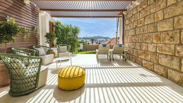Une terrasse extérieure couverte, aménagée avec des meubles en osier, un pouf jaune et offrant une vue sur la ville en arrière-plan.
