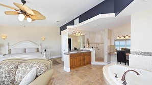 A spacious bedroom with a large bed, a sitting area with a sofa, a separate bathroom with a bathtub, and a ceiling fan.