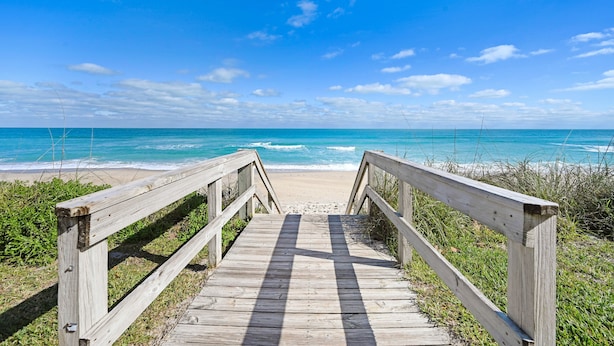 A wooden boardwalk leading to a beach with clear blue water and a bright sky.
