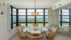 Ein Essbereich mit einem Glastisch, Korbstühlen und Blick auf das Meer durch große Fenster.