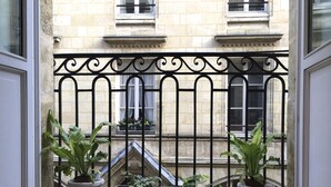 Une vue à travers une fenêtre, avec un balcon en fer forgé, des plantes en pot et la façade d’un immeuble ornée de fenêtres et de pierres apparentes.