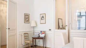 A room with a wooden floor, a radiator, a chair, a table with books, and two framed pictures on the wall.