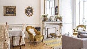 Une pièce avec une cheminée, deux fauteuils, une table recouverte d’une nappe à carreaux et un miroir au-dessus de la cheminée.