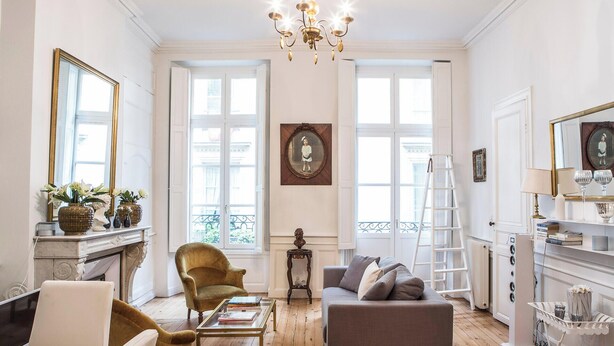 Un salon bien éclairé, doté d’une cheminée, d’un lustre et offrant une vue dégagée grâce à de grandes fenêtres.