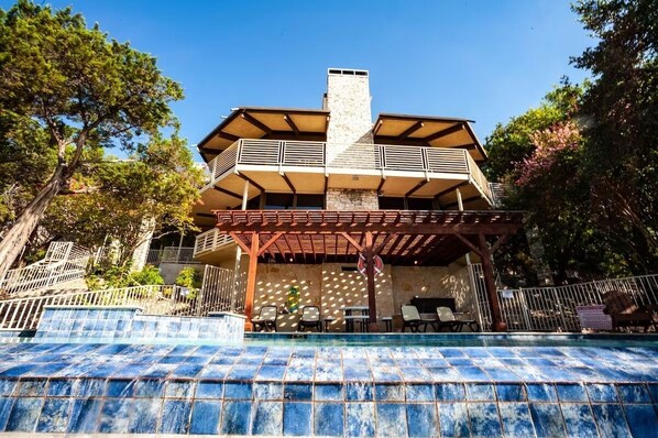 A two-story building with a stone chimney, a covered patio area with outdoor seating, and a swimming pool.