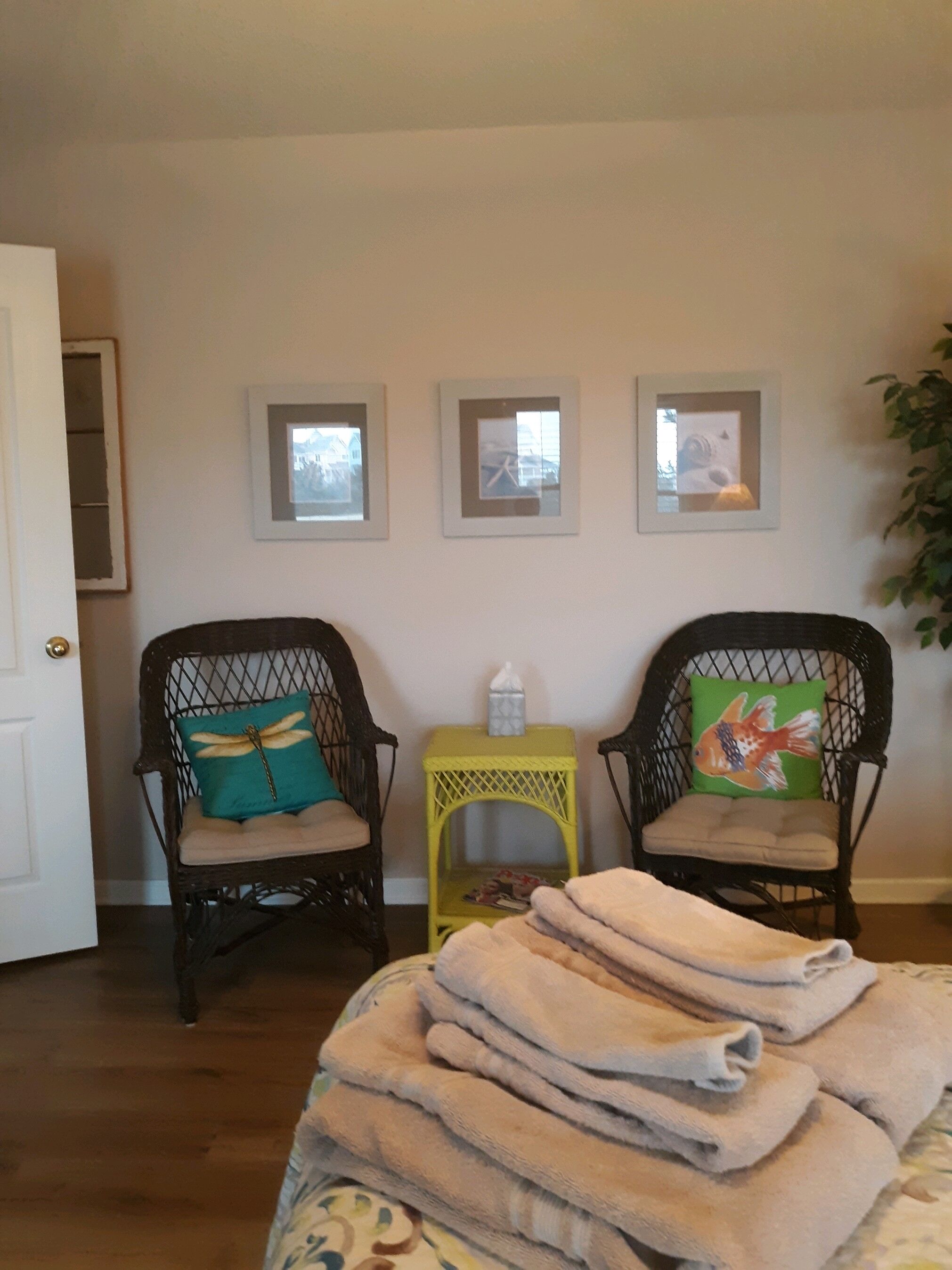 A room with two wicker chairs, a small yellow side table, and a stack of folded towels on a bed.