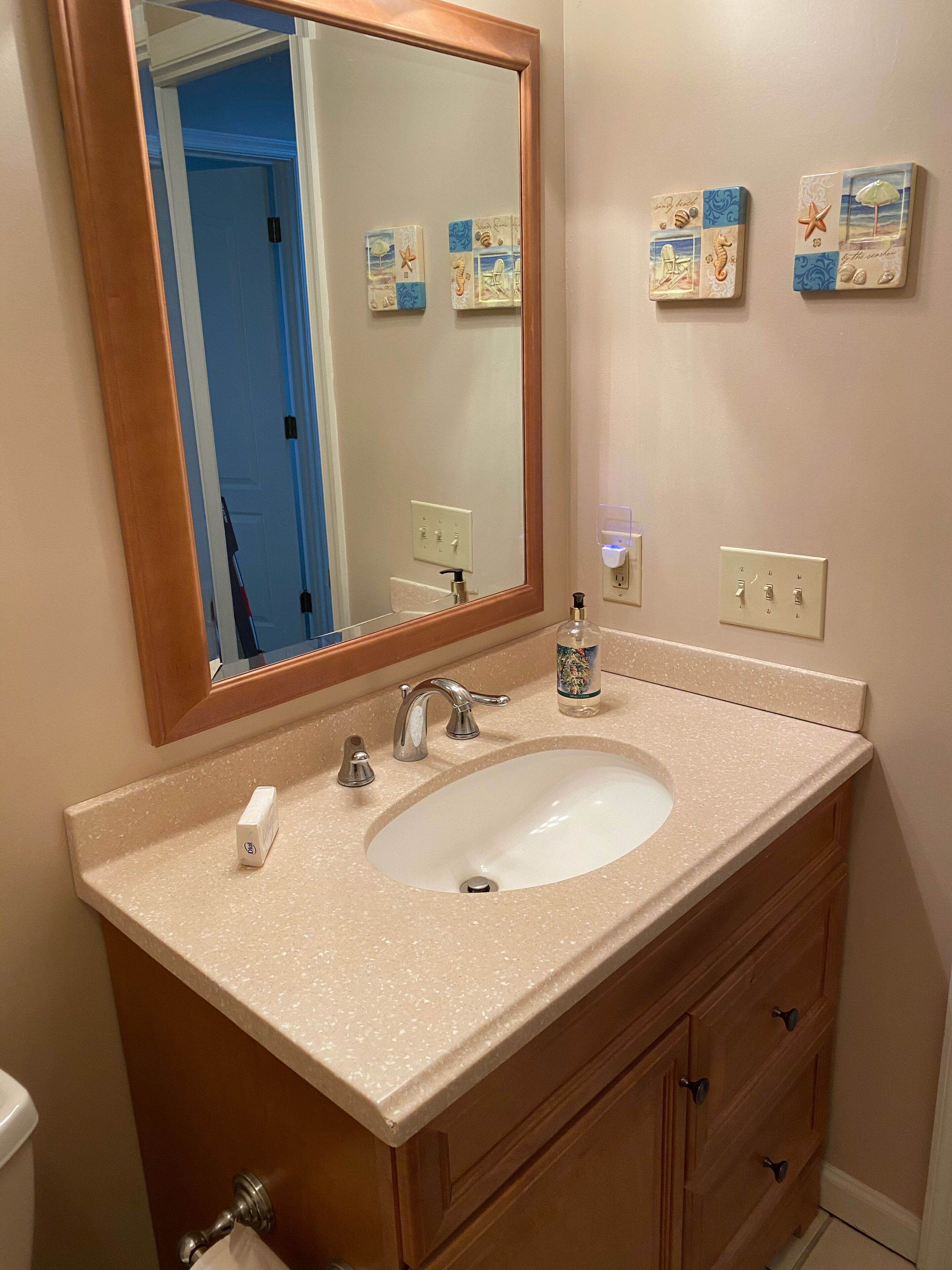 Un baño con encimera color beige, lavabo blanco, mueble de tocador de madera y un espejo. Hay dos interruptores de luz en la pared y un dispensador de jabón sobre la encimera.