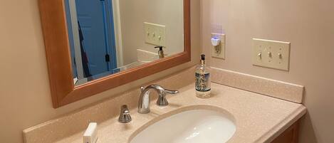 Un baño con encimera color beige, lavabo blanco, mueble de tocador de madera y un espejo. Hay dos interruptores de luz en la pared y un dispensador de jabón sobre la encimera.