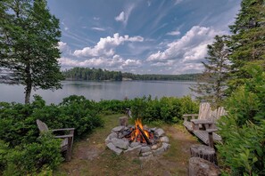 Un camping a orillas del lago con un hogar, sillas de madera y vistas al lago y al bosque.