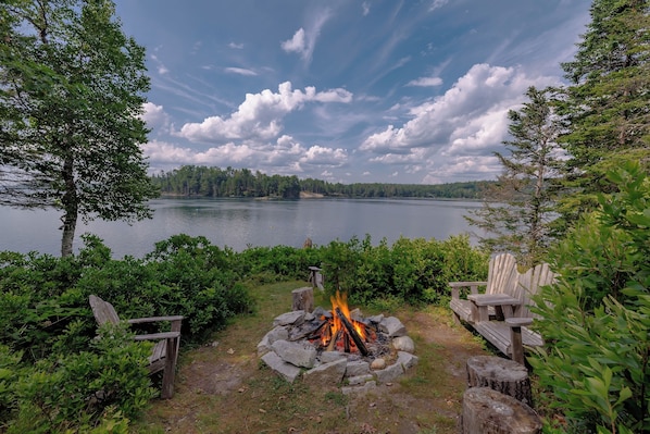 Un camping a orillas del lago con un hogar, sillas de madera y vistas al lago y al bosque.