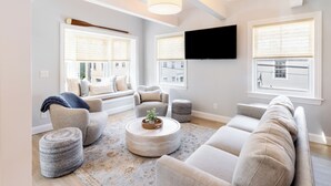 A living room with a sectional sofa, round ottomans, a coffee table, and a TV mounted on the wall. - Condo w/Spiral Staircase to Loft, Perfect for Kids (Newport)