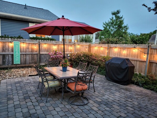 A patio with a table and chairs, a red umbrella, and a wooden fence with string lights.