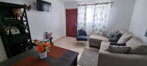 Ein Wohnzimmer mit einer Couch, einem Sessel, einem Couchtisch, einem Fernseher und einer Tür mit Vorhängen.