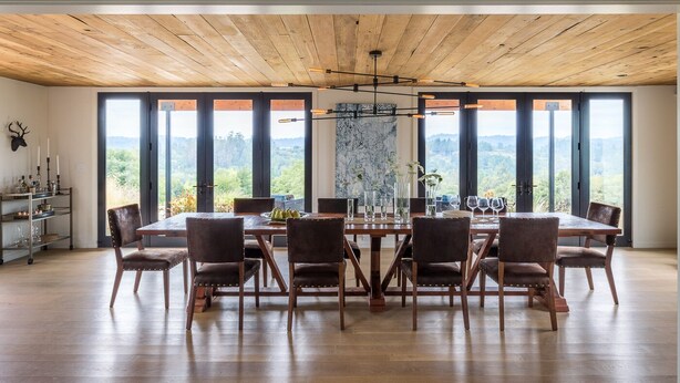 Une salle à manger avec une grande table en bois, huit chaises et une vue sur un paysage montagneux, visible à travers de grandes fenêtres.