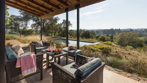 Un espace extérieur aménagé, abrité et doté de mobilier en osier, avec une table où sont disposés des fruits, et offrant une vue sur une piscine et un paysage verdoyant.