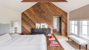 Une chambre avec un plafond en pente, un lit, une commode, un banc et une grande fenêtre.