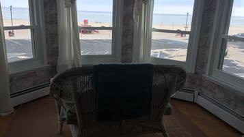 A room with a wicker chair, a view of the beach, and a parking lot.